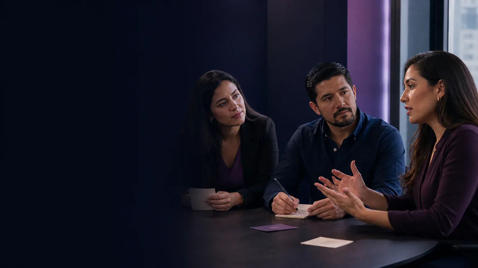 Three Latin American professionals practice an assertive conversation while listening, explaining, and taking notes.