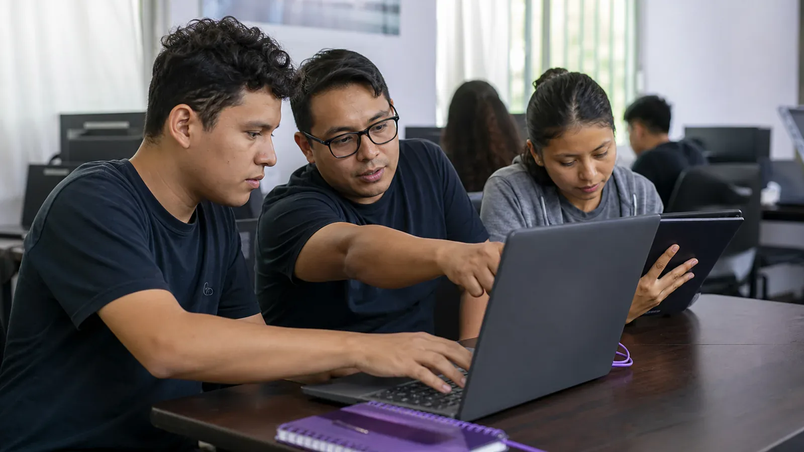 A mentor and two learners work together on a practical digital project using a laptop and tablet.