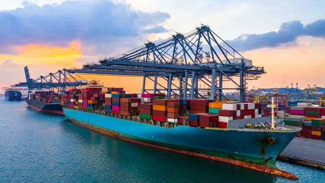 Container ships at a dock at sunset, with port cranes and an orange and blue sky.