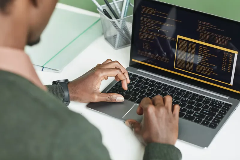 African American man coding on a laptop with code on screen, seen from over the shoulder.