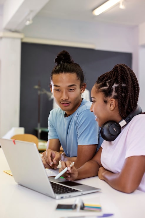 Two diverse students collaborate on a laptop, one with a top knot and one with braids and headphones.