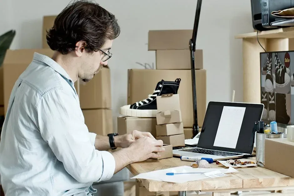 Man packing small boxes at a desk with a laptop, surrounded by shipping boxes and a sneaker display, indicating an e-commerce business.