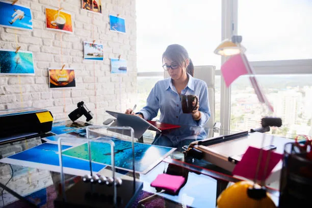 Woman working at a glass desk with tablet, coffee mug, and photos on white brick wall.