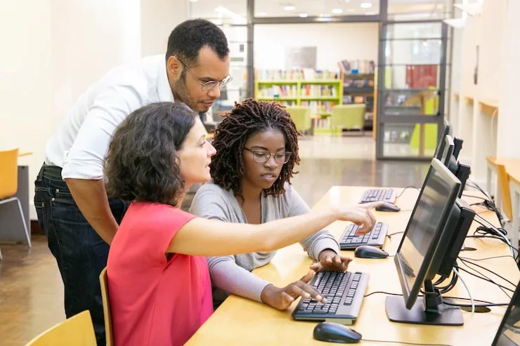 Three people collaborate in a computer lab, one woman points at the screen while another types.