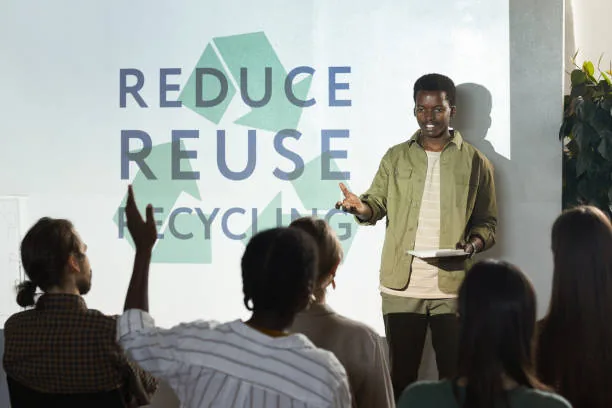 Black man presents "Reduce, Reuse, Recycle" to an attentive audience, with one person raising a hand.