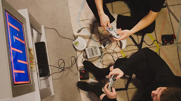 Two people play retro video games on the floor with consoles and controllers, overhead view.