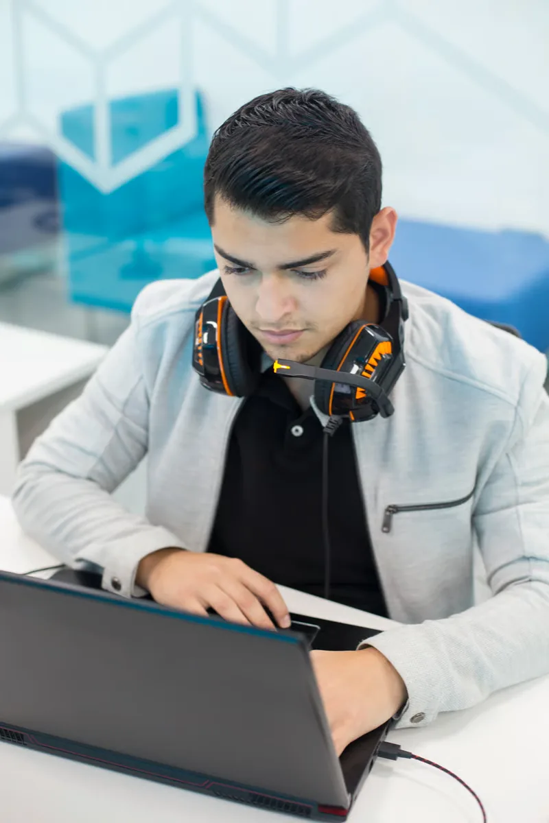 Focused young man with gaming headphones around his neck, using a laptop in a modern setting.