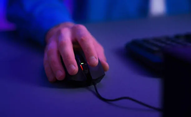 Gamer's hand using a gaming mouse with LED lights on a dark desk with a keyboard.