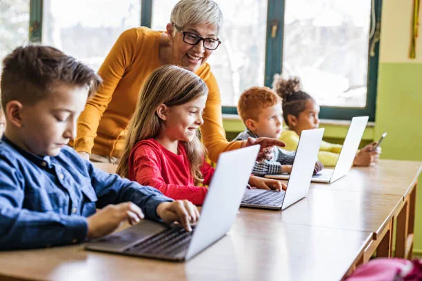 Smiling teacher helps elementary kids with laptops in a modern classroom, fostering digital learning.