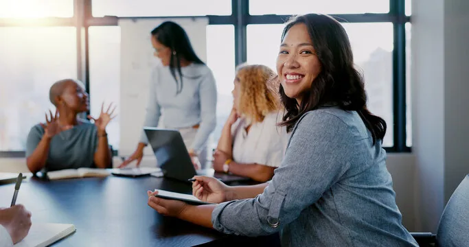 Smiling woman in foreground with tablet, diverse team of women in business meeting in background.