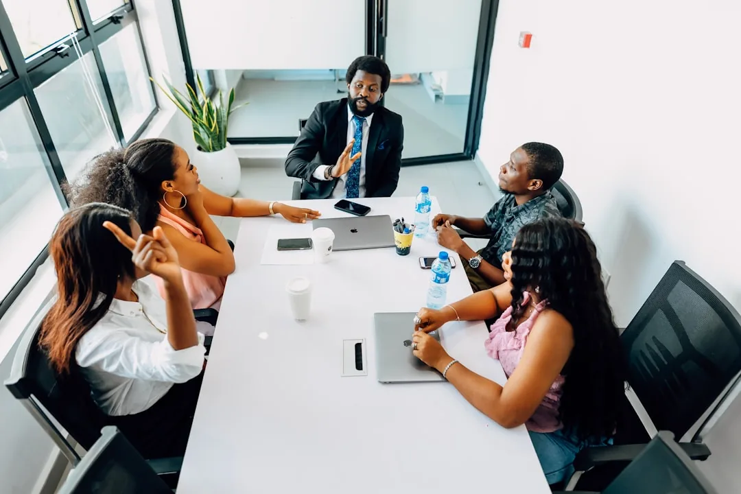 Five diverse professionals in a modern office meeting. A man leads the discussion, while others listen attentively and one woman points.