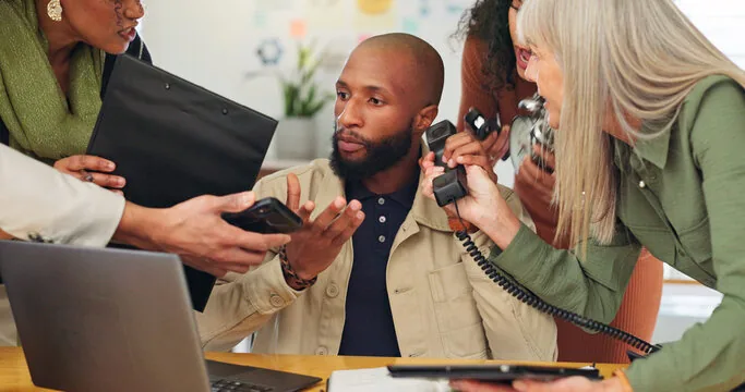 Businessman overwhelmed by colleagues handing him documents, a phone, and a mobile.