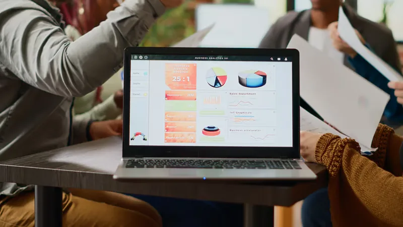 Laptop displaying a business analytics dashboard with charts and data, with people in the background.
