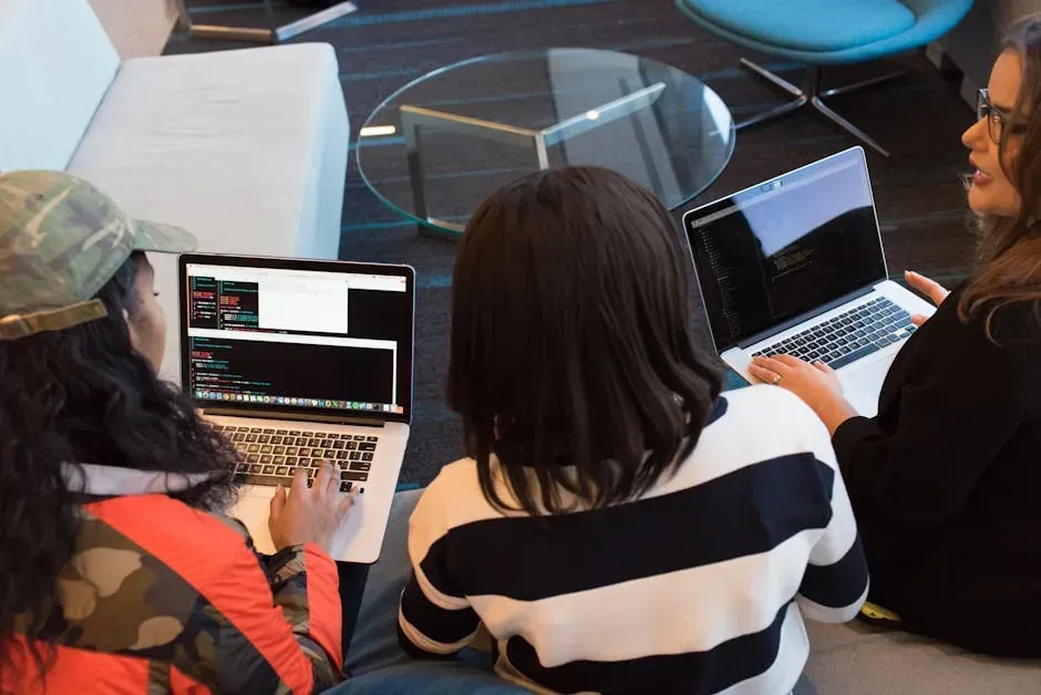 Three women coding on laptops with code on screen, collaborating in a modern office space.