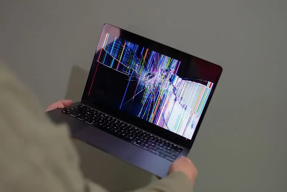 Person holding a laptop with a broken LCD screen, showing cracks with bright colorful lines and distorted pixels.