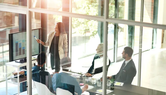Pregnant woman presents bar graphs on a screen to colleagues in a glass-walled meeting room.