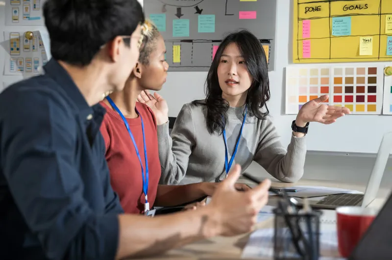 Three diverse colleagues in a design meeting, an Asian woman explains with gestures.