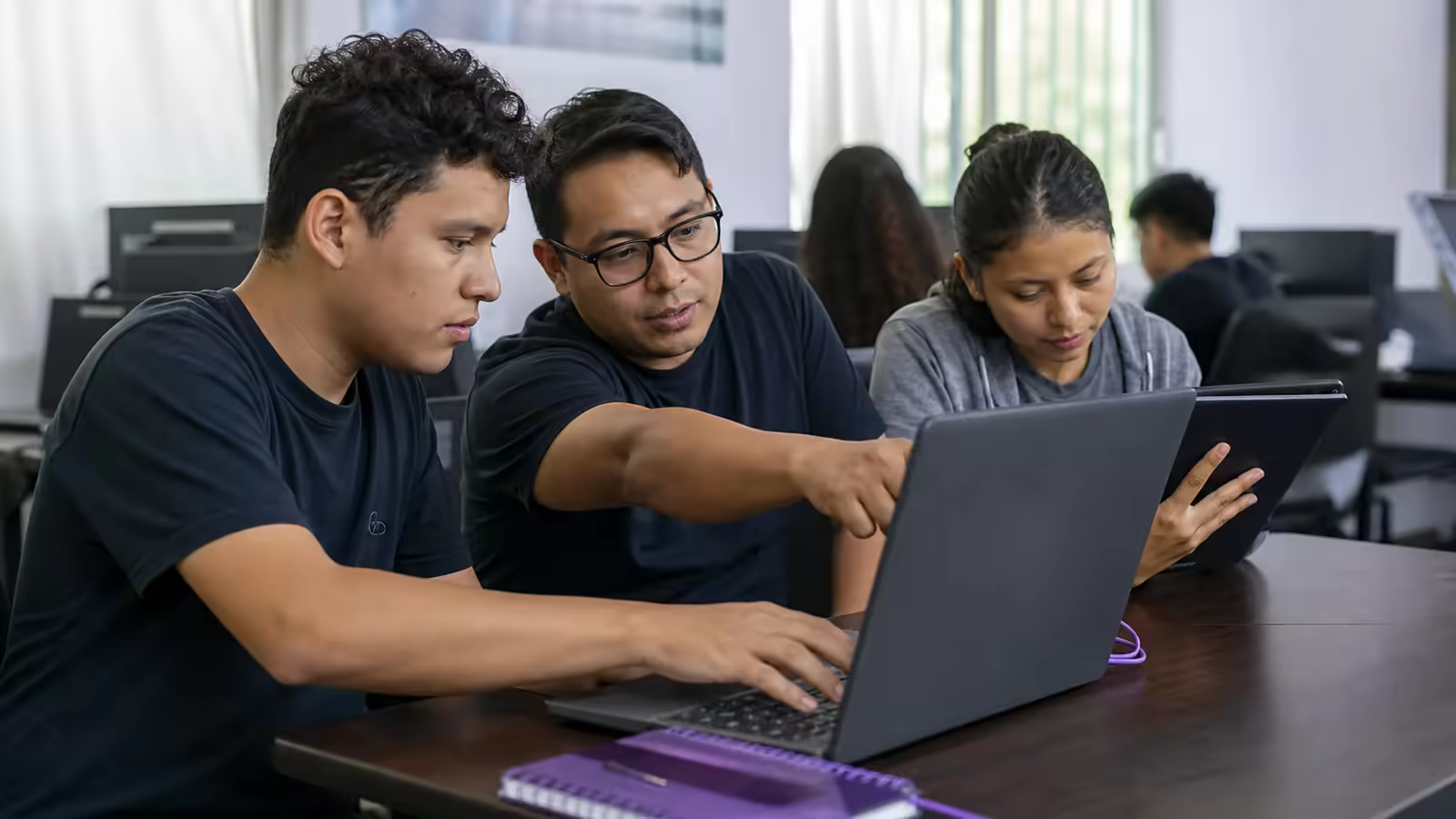 Un mentor y dos estudiantes trabajan juntos en un proyecto digital práctico con una laptop y una tableta.